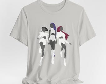 May include: A light grey t-shirt with a graphic of three stylized figures in motion. The figures are in white, black, and purple, with varying hair colours. The design is centered on the shirt.