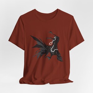 May include: A rust-colored t-shirt featuring a black graphic of a cloaked figure with a chain around its neck. The figure has a skeletal face and is set against a dark background. The t-shirt is made of a soft material.