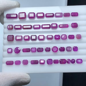 May include: A collection of various-shaped, faceted ruby gemstones. The gemstones are arranged on a white tray, showcasing a range of cuts including emerald, square, and oval shapes. The rubies display a deep red-pink hue.