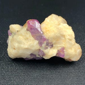 Könnte beinhalten: Natürliches Mineralpräparat mit rosavioletten Kristallen, eingebettet in eine cremeweiße Matrix. Die Kristalle sind durchscheinend, und die Matrix hat eine raue, strukturierte Oberfläche. Naturformation.