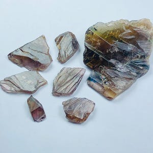 Könnte beinhalten: Eine Sammlung roher, durchscheinender Fluoritkristalle in verschiedenen Formen und Größen. Die Kristalle zeigen eine Reihe von Farben, darunter Lila, Braun und Gelb, mit sichtbaren inneren Streifen. Die Anordnung befindet sich auf weißem Hintergrund.