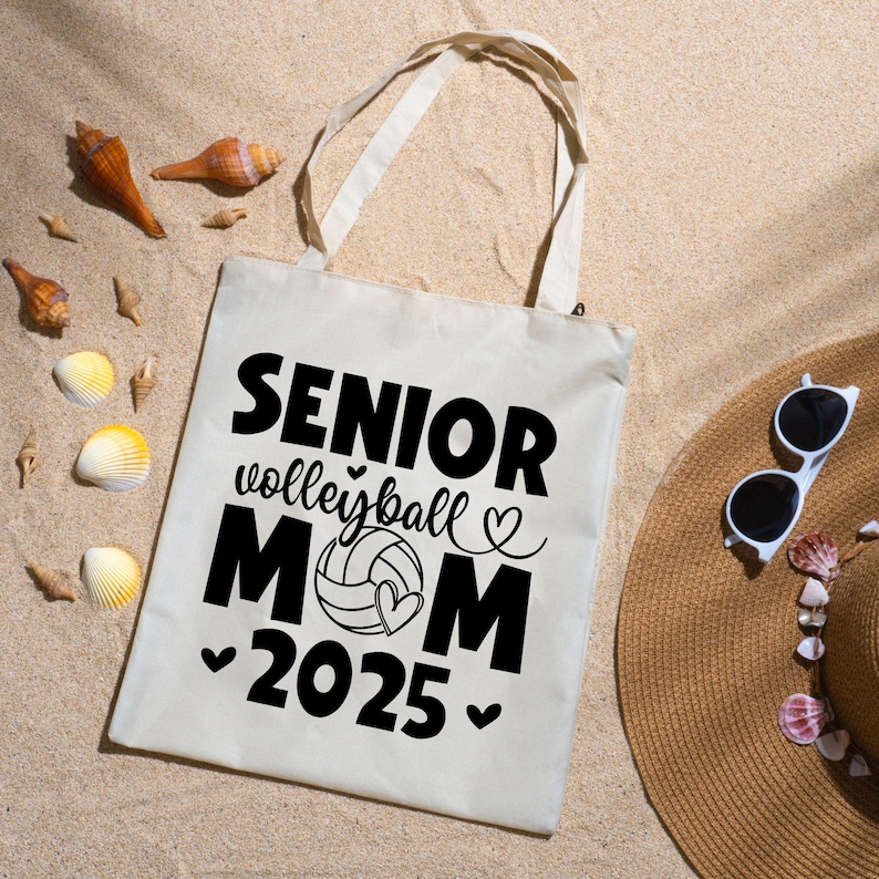 May include: Cream-colored tote bag with black text that reads "SENIOR volleyball MOM 2025" with a volleyball and heart graphic. The bag has two handles and is on a sandy surface with seashells and a straw hat.