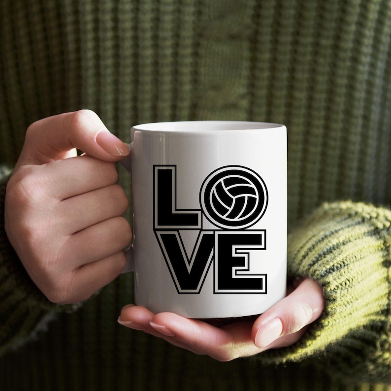 May include: White ceramic mug with the word "LOVE" in bold black letters, with a volleyball graphic replacing the "O". The mug is held by a person wearing a green sweater. A great gift for volleyball enthusiasts.