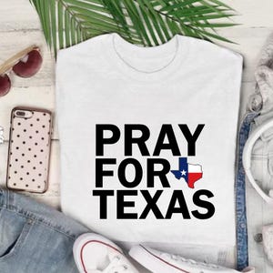 K&ouml;nnte beinhalten: Wei&szlig;es T-Shirt mit dem Text "PRAY FOR TEXAS" in gro&szlig;en schwarzen Buchstaben. Das Wort "FOR" enth&auml;lt eine Texas-Umrisslinie mit einem blauen Stern und roten und wei&szlig;en Akzenten. Das Shirt wird mit Sonnenbrille, Telefon und Jeans gezeigt.