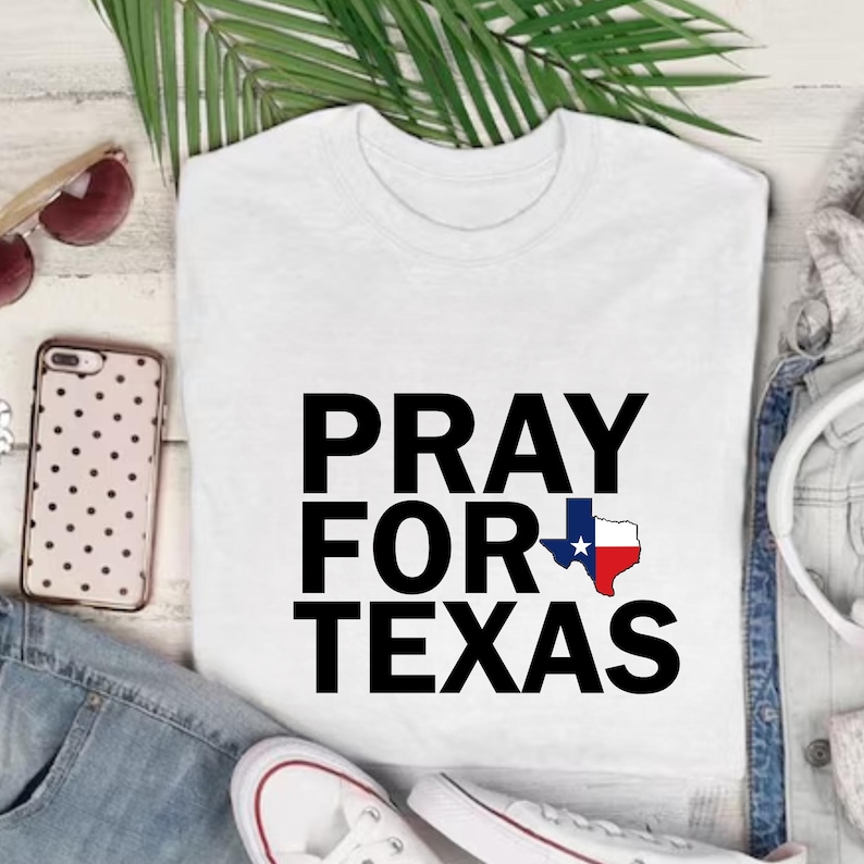 K&ouml;nnte beinhalten: Wei&szlig;es T-Shirt mit dem Text "PRAY FOR TEXAS" in gro&szlig;en schwarzen Buchstaben. Das Wort "FOR" enth&auml;lt eine Texas-Umrisslinie mit einem blauen Stern und roten und wei&szlig;en Akzenten. Das Shirt wird mit Sonnenbrille, Telefon und Jeans gezeigt.