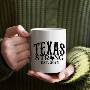 K&ouml;nnte beinhalten: Wei&szlig;e Keramik-Tasse mit dem Schriftzug "TEXAS STRONG" in schwarzer, fetter Schrift. Darunter der Umriss des Bundesstaates Texas und der Text "EST. 2025". Die Tasse wird von einer Person gehalten, die einen gr&uuml;nen Pullover tr&auml;gt.