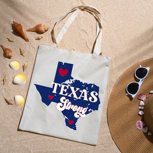 K&ouml;nnte beinhalten: Cremefarbene Tragetasche mit blauem Texas-Umriss, roten Herzen und dem Schriftzug "TEXAS Strong" in wei&szlig;. Die Tasche liegt auf Sand, mit Muscheln und einem Strohhut mit Sonnenbrille.