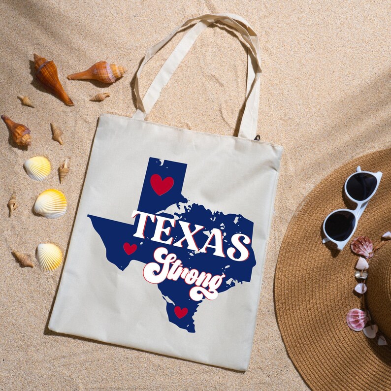 K&ouml;nnte beinhalten: Cremefarbene Tragetasche mit blauem Texas-Umriss, roten Herzen und dem Schriftzug "TEXAS Strong" in wei&szlig;. Die Tasche liegt auf Sand, mit Muscheln und einem Strohhut mit Sonnenbrille.