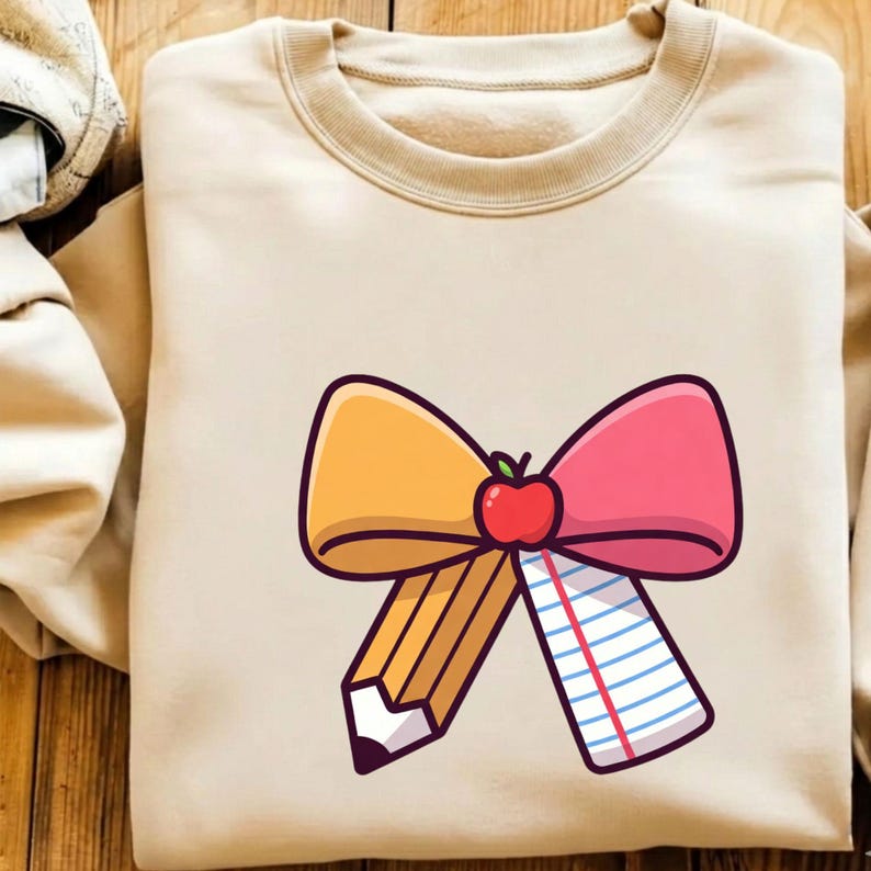 May include: A beige sweatshirt featuring a colorful graphic of a bow made of a yellow pencil, pink and yellow bow, and a piece of lined paper. A red apple sits in the center of the bow. The sweatshirt has a crew neck and long sleeves.