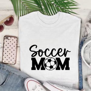 Pu&ograve; includere: Maglietta bianca con la scritta nera "Soccer MOM" e un pallone da calcio. La maglietta &egrave; esposta con occhiali da sole, una custodia per telefono e scarpe da ginnastica bianche, su una superficie bianca.