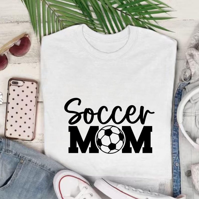 Pu&ograve; includere: Maglietta bianca con la scritta nera "Soccer MOM" e un pallone da calcio. La maglietta &egrave; esposta con occhiali da sole, una custodia per telefono e scarpe da ginnastica bianche, su una superficie bianca.