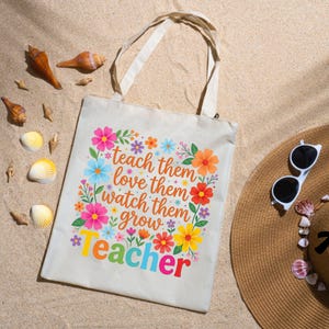 May include: A beige canvas tote bag with the words "teach them, love them, watch them grow, teacher" in colorful text and floral accents. The bag is on a sandy surface with seashells and a straw hat with sunglasses.