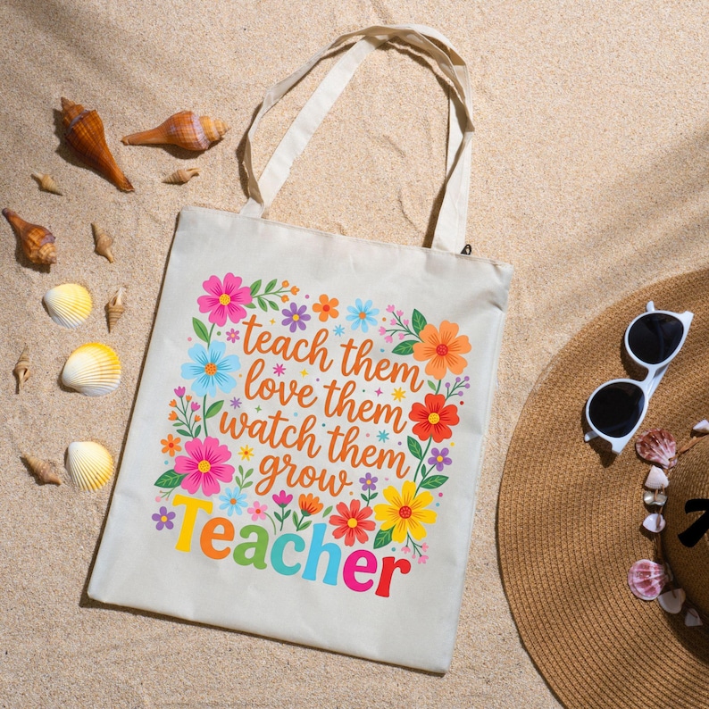 May include: A beige canvas tote bag with the words "teach them, love them, watch them grow, teacher" in colorful text and floral accents. The bag is on a sandy surface with seashells and a straw hat with sunglasses.