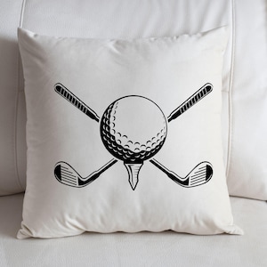 May include: A white square pillow with a black graphic of a golf ball on a tee, crossed golf clubs. The golf ball is in the center, with the clubs behind it. The pillow is on a white surface.