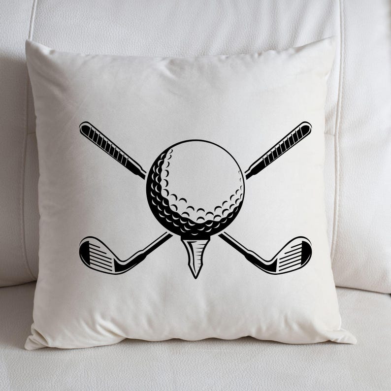 May include: A white square pillow with a black graphic of a golf ball on a tee, crossed golf clubs. The golf ball is in the center, with the clubs behind it. The pillow is on a white surface.