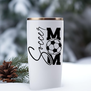 Pu&ograve; includere: Bicchiere termico bianco con bordo dorato. Il bicchiere presenta le parole "Soccer Mom" in corsivo nero e lettere maiuscole, con una grafica di pallone da calcio. Lo sfondo &egrave; una scena esterna innevata.