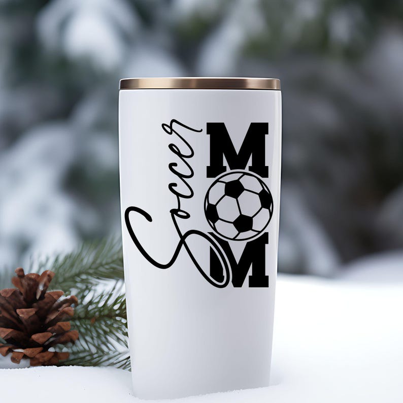 Pu&ograve; includere: Bicchiere termico bianco con bordo dorato. Il bicchiere presenta le parole "Soccer Mom" in corsivo nero e lettere maiuscole, con una grafica di pallone da calcio. Lo sfondo &egrave; una scena esterna innevata.