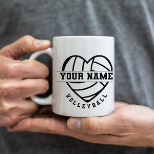 Op de afbeelding: Witte keramische mok met een zwart hartvormig volleybalontwerp. Het ontwerp bevat de tekst "YOUR NAME" boven een horizontale lijn en het woord "VOLLEYBALL" eronder. De mok wordt door een persoon vastgehouden.
