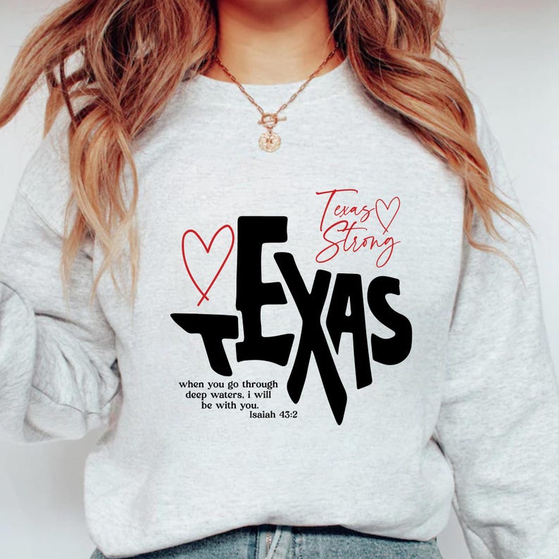 K&ouml;nnte beinhalten: Ein hellgraues Sweatshirt mit dem Wort "TEXAS" in gro&szlig;en schwarzen Buchstaben, einem roten Herz und den Worten "Texas Strong" in roter Schrift. Das Sweatshirt hat auch den Text "when you go through deep waters. I will be with you. Isaiah 43:2."