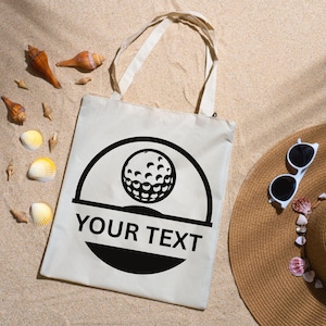 May include: A cream-colored tote bag with a black golf ball design and the text "YOUR TEXT". The bag has two handles and is lying on sand, next to a straw hat and sunglasses. Seashells are scattered around the bag.