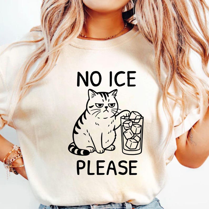 May include: Cream-colored t-shirt with a black graphic of a grumpy cat and a glass of ice cubes. The text "NO ICE PLEASE" is printed above and below the cat illustration.