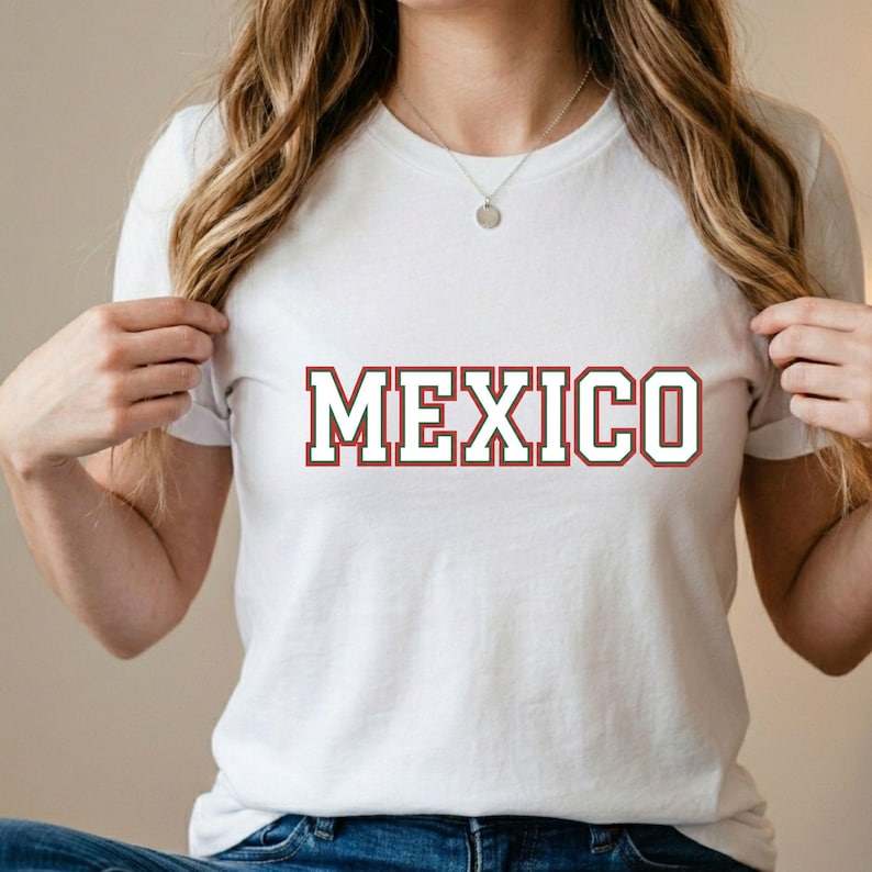May include: A white short-sleeved t-shirt featuring the word MEXICO printed across the chest in a bold, collegiate-style font with red lettering and a thin green outline. The shirt has a classic crew neckline and a simple, clean design.