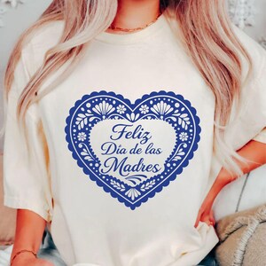 May include: Cream-colored t-shirt featuring a blue heart-shaped design with floral and dot patterns. The heart contains the text "Feliz D&iacute;a de las Madres" in a script font, celebrating Mother's Day.