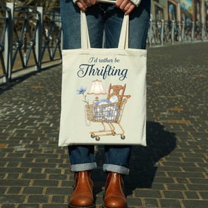 Może przedstawiać: Kremowa torba typu tote z napisem "I'd rather be Thrifting". Torba przedstawia grafikę w&oacute;zka sklepowego wypełnionego przedmiotami w stylu vintage, w tym lampą, krzesłem oraz niebiesko-białą porcelaną. Torba jest trzymana przez osobę ubraną w niebieskie dżinsy i brązowe buty.