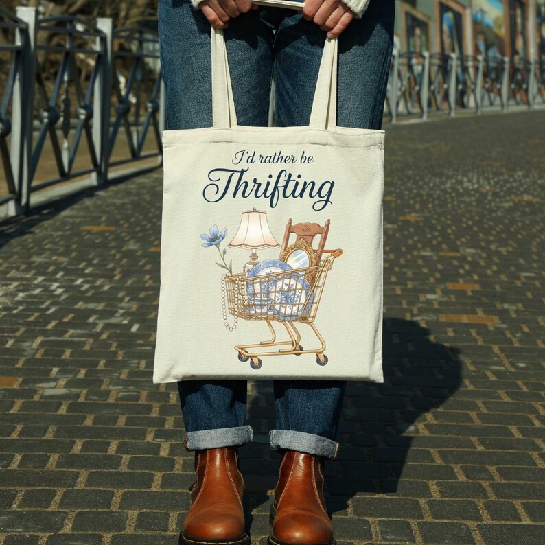 Może przedstawiać: Kremowa torba typu tote z napisem "I'd rather be Thrifting". Torba przedstawia grafikę w&oacute;zka sklepowego wypełnionego przedmiotami w stylu vintage, w tym lampą, krzesłem oraz niebiesko-białą porcelaną. Torba jest trzymana przez osobę ubraną w niebieskie dżinsy i brązowe buty.