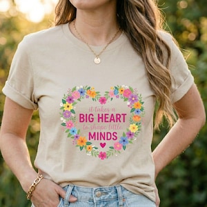 May include: Beige t-shirt with a floral heart design. The heart is composed of colorful flowers and contains the text "it takes a BIG HEART to shape little MINDS". A gold necklace and bracelet are also visible.