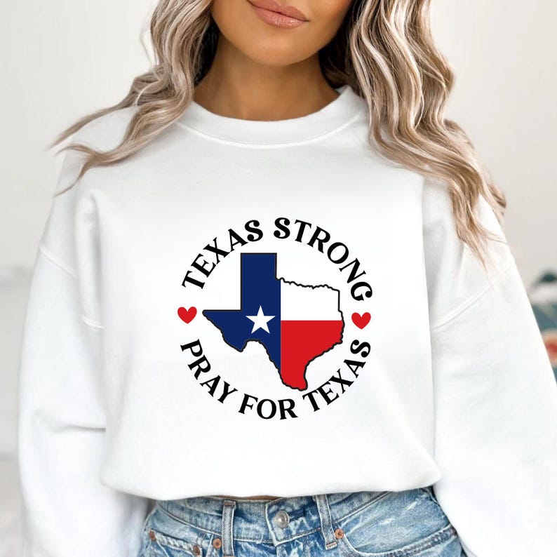 K&ouml;nnte beinhalten: Wei&szlig;es Sweatshirt mit einem kreisf&ouml;rmigen Design, das die Flagge des Bundesstaates Texas und die Worte "Texas Strong" und "Pray for Texas" zeigt. Die Flagge ist blau, wei&szlig; und rot, mit einem wei&szlig;en Stern. Zwei rote Herzen befinden sich auf beiden Seiten des Designs.