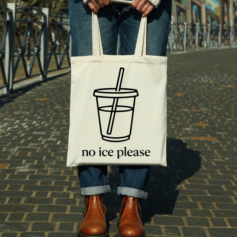 May include: A cream-colored tote bag with a black graphic of a beverage cup with a straw. The text "no ice please" is printed below the cup. The bag is held by a person wearing blue jeans and brown boots.