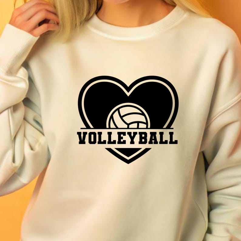 Op de afbeelding: Witte sweatshirt met een zwart hartontwerp met een volleybal erin. Het woord "VOLLEYBALL" is in vette, zwarte letters onder het hart gedrukt. Een casual, comfortabele stijl.