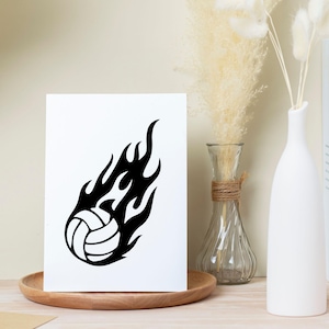 Op de afbeelding: Een zwart-wit illustratie van een volleybal omringd door vlammen. Het ontwerp staat op een witte kaart, gepresenteerd op een houten dienblad. De afbeelding heeft een sportthema.