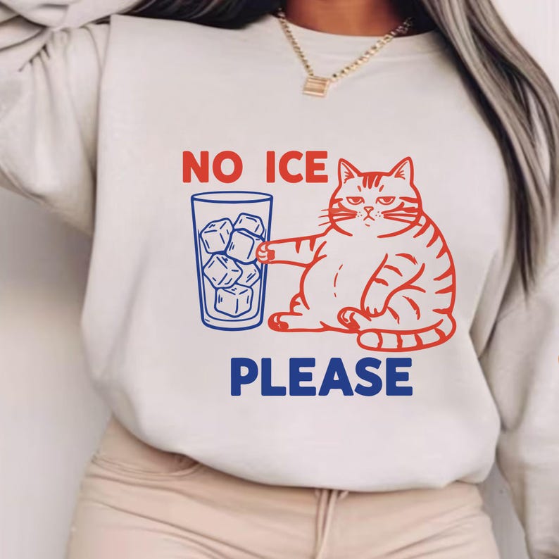 May include: Cream-colored sweatshirt featuring a red and white cartoon cat holding a blue glass filled with ice cubes. The text "NO ICE PLEASE" is printed above and below the cat illustration in red and blue.