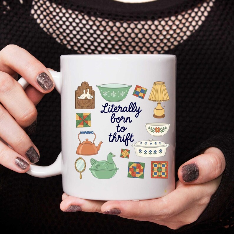 Może przedstawiać: Biały ceramiczny kubek z napisem "Literally born to thrift" i r&oacute;żnymi ilustracjami w stylu vintage. Ilustracje obejmują lampę, czajnik i dekoracyjne miski. Kubek jest trzymany przez osobę.