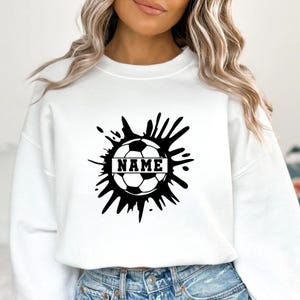 Pu&ograve; includere: Felpa bianca con un design a pallone da calcio nero e la parola "NAME" in un riquadro rettangolare. Il pallone da calcio &egrave; circondato da un design a spruzzo nero. Uno stile casual e sportivo.