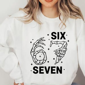 May include: White sweatshirt with a black graphic design. The design features two skeleton hands forming the numbers six and seven, with the words "SIX" and "SEVEN" printed below. The design includes small star accents.