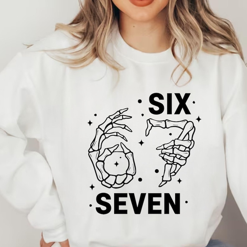 May include: White sweatshirt with a black graphic design. The design features two skeleton hands forming the numbers six and seven, with the words "SIX" and "SEVEN" printed below. The design includes small star accents.