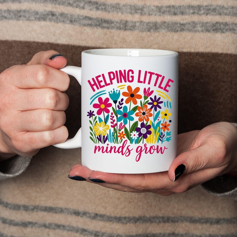May include: White ceramic mug with the text "HELPING LITTLE minds grow" in pink and teal. The mug features a colorful floral design with orange, blue, yellow, and purple flowers. The mug is held by a person.