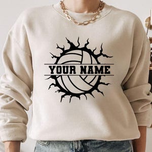 Op de afbeelding: Beige sweatshirt met een zwarte volleybalafbeelding. Het volleybalontwerp is omgeven door een gekarteld barsteffect, met de tekst "YOUR NAME" in een vetgedrukte, blokletter. De sweatshirt heeft een ronde hals en lange mouwen.