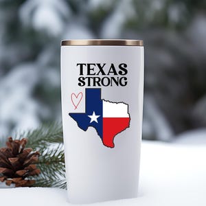 K&ouml;nnte beinhalten: Wei&szlig;er Reisebecher mit goldenem Rand, mit dem Text "TEXAS STRONG" &uuml;ber einer Grafik der Texas-Staatsflagge und einem roten Herzen. Der Becher ist f&uuml;r hei&szlig;e oder kalte Getr&auml;nke konzipiert.