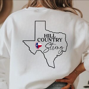 K&ouml;nnte beinhalten: Wei&szlig;es Sweatshirt mit schwarzer Umrisslinie des Bundesstaates Texas. Die Worte "HILL COUNTRY" stehen dar&uuml;ber und "Strong" in kursiver Schrift darunter. Ein Herz mit dem Design der Texas-Flagge befindet sich innerhalb der Texas-Umrisslinie.