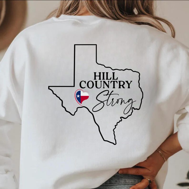 K&ouml;nnte beinhalten: Wei&szlig;es Sweatshirt mit schwarzer Umrisslinie des Bundesstaates Texas. Die Worte "HILL COUNTRY" stehen dar&uuml;ber und "Strong" in kursiver Schrift darunter. Ein Herz mit dem Design der Texas-Flagge befindet sich innerhalb der Texas-Umrisslinie.