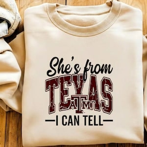 May include: A beige sweatshirt with a crew neck and long sleeves. The front features the text "She's from TEXAS A&M - I CAN TELL" in black and maroon lettering. The sweatshirt is made of a soft material.