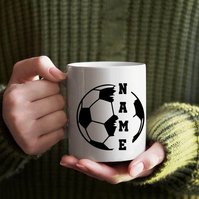Pu&ograve; includere: Tazza in ceramica bianca con un disegno di pallone da calcio nero e la parola "NAME" in un carattere verticale. La tazza &egrave; tenuta da una persona che indossa un maglione verde.