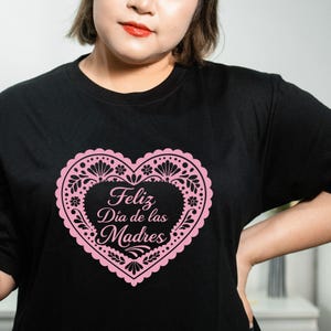 May include: Black t-shirt featuring a pink heart-shaped design with floral and decorative elements. The heart contains the text "Feliz D&iacute;a de las Madres" in elegant script, perfect for Mother's Day.