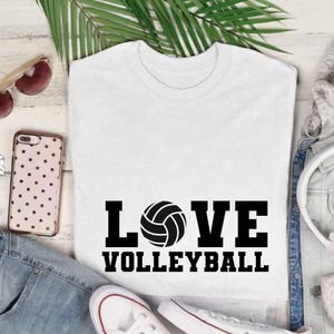 May include: White t-shirt with the words "LOVE VOLLEYBALL" in black block letters and a volleyball graphic. The shirt is displayed with sunglasses, a phone, and a pair of white sneakers.