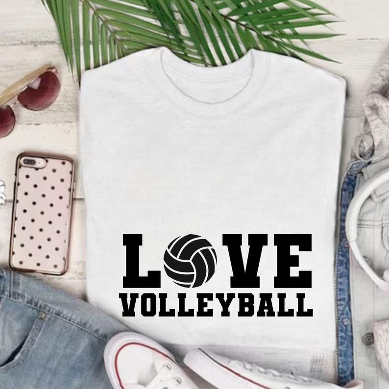 May include: White t-shirt with the words "LOVE VOLLEYBALL" in black block letters and a volleyball graphic. The shirt is displayed with sunglasses, a phone, and a pair of white sneakers.
