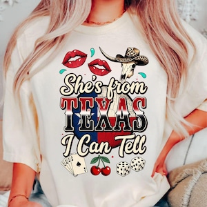 May include: Cream-colored t-shirt with a graphic that reads "She's from TEXAS I Can Tell." The design includes red lips, a steer skull with a cowboy hat, playing cards, cherries, and dice.